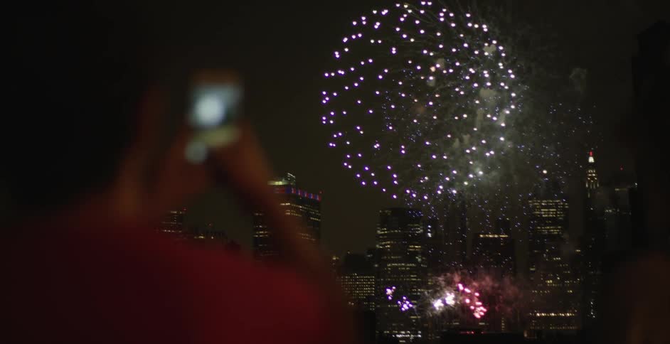 A person uses a phone to photographic fireworks over a city at night