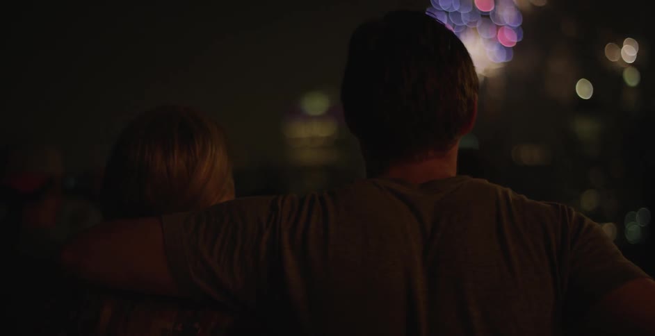 A man puts his arm around a woman then kisses her forehead while they watch fireworks at night