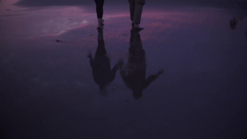 Two people are walking in the water by the shore at sunset
