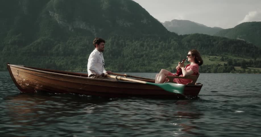 Man rowing a wooden canoe on a lake while his girlfriend is sitting in front of him playing a string instrument