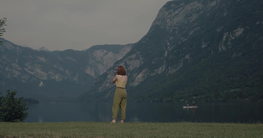 A photographer raises her camera to take a photograph of the landscape while standing on the shore of a lake
