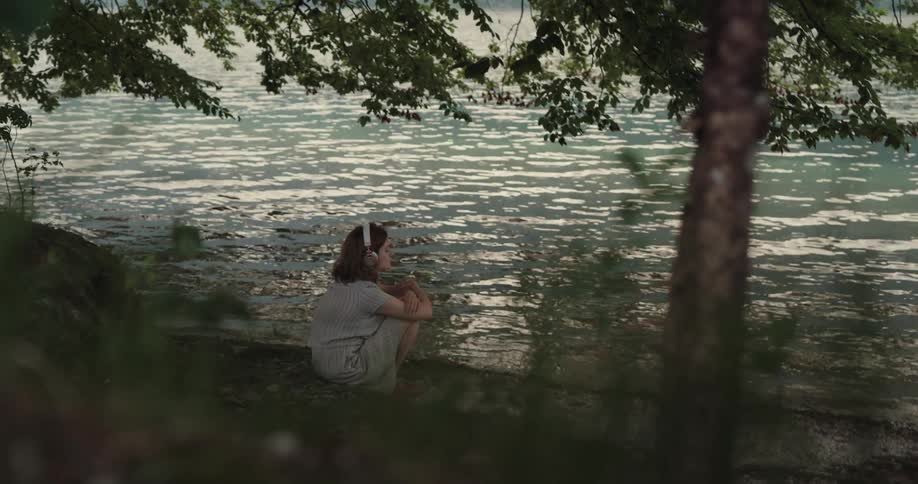 A woman wears headphones while sitting near a lake