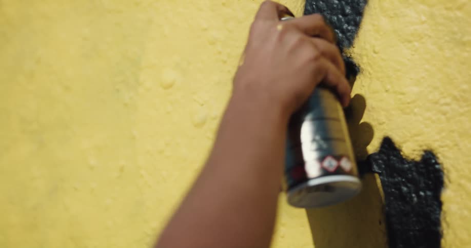 A hand holding a spray paint can paints black on a yellow wall in the sunshine