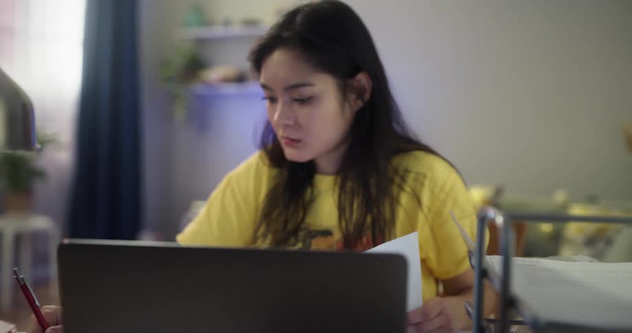 Teenage asian girl sitting at the desk in the living room studying on her laptop and notebook