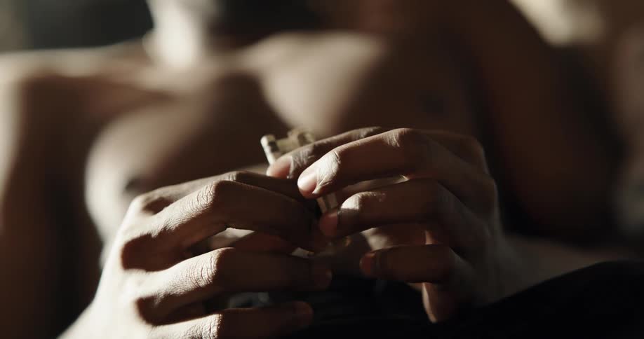 A shirtless man tightly rolls up a 10 dollar bill while reclining indoors