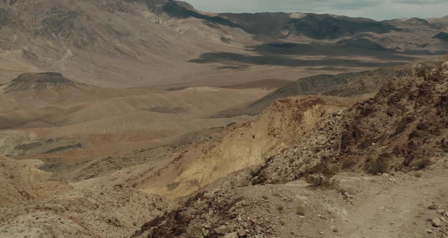 A jeep sits on top of a desert mountain road with a valley below and mountains in the distance