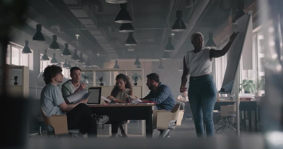 Black woman standing next to a white board leading a meeting in an office space shouting at the employees