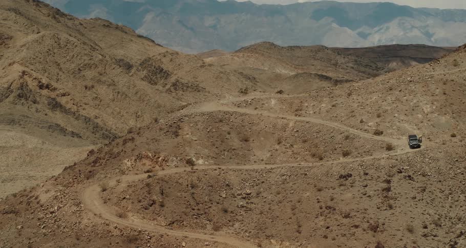 A jeep is parked high on a winding desert mountain road with mountains in the distance and a man is walking around the jeep