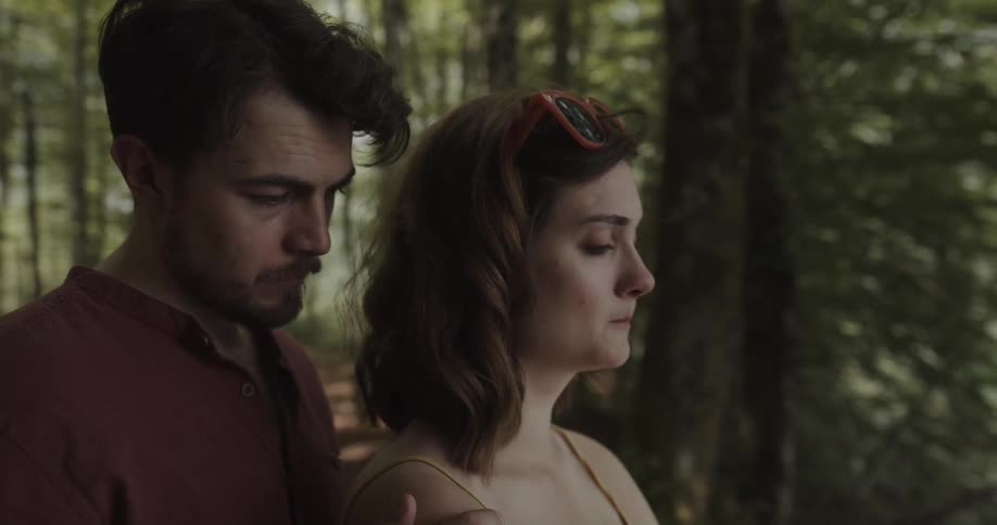 Upset woman standing outdoors in a forest area while her boyfriend approaches from behind and hugs her