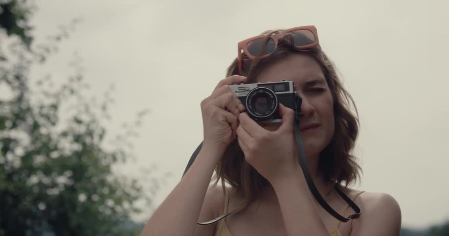 A woman raises a camera to her face to take a photograph while standing outdoors on a cloudy day
