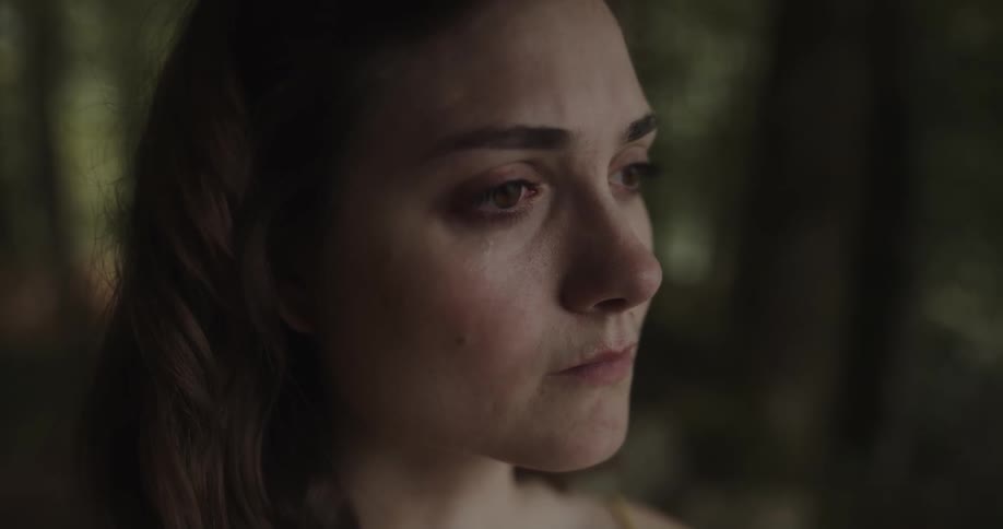 A woman standing in a shady grove looks out with a sad expression on her face