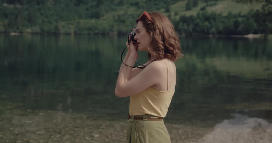 White woman standing near the water of a lake taking pictures of the area with her digital camera during the day