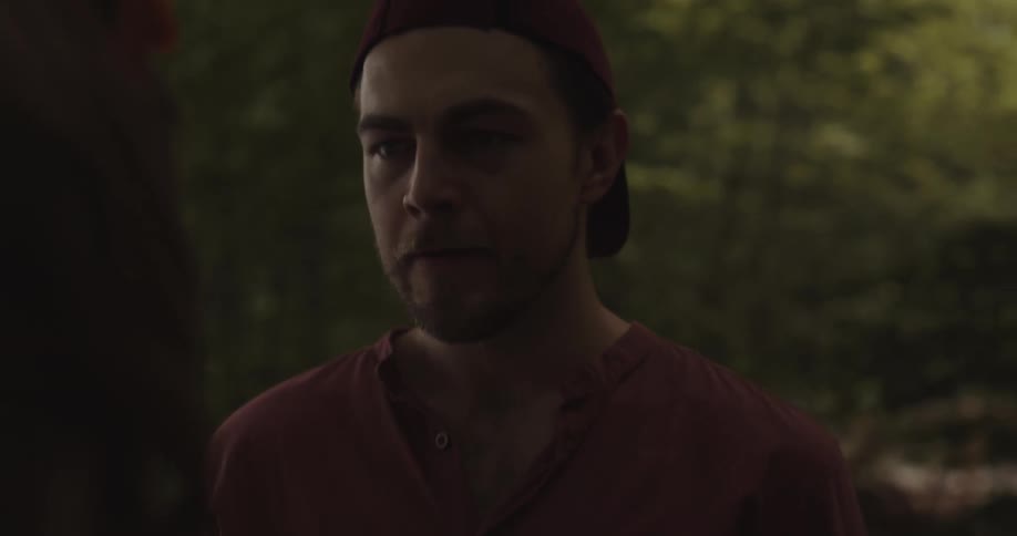 Man wearing a hat backwards and a woman argue while standing in a forest next to a lake