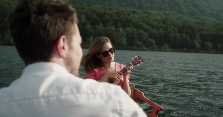 Man and woman sitting in a wooden canoe in the middle of the lake while she is happily playing a mini guitar and he is rowing