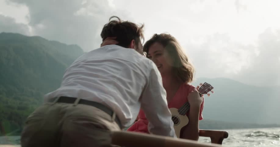 Man moves across bobbing rowboat near misty mountainous shore to kiss woman who was playing ukulele