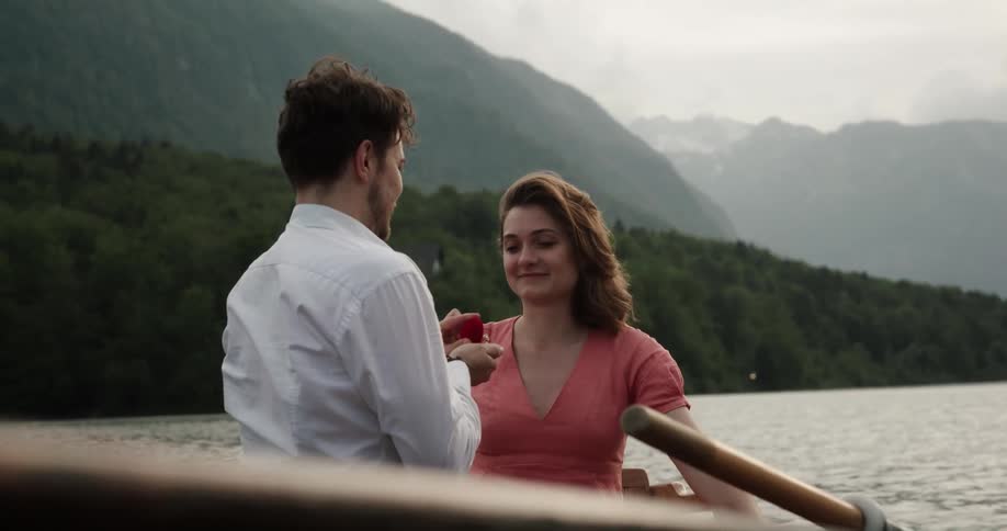 Man proposes to woman while they sit in rowboat near forest covered shore with misty mountains in the background