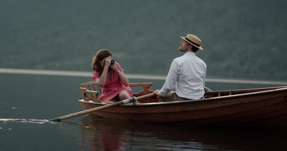 Man and woman sitting on a small wooden boat in the middle of a lake while the woman is taking pictures of the man who is posing with a hat