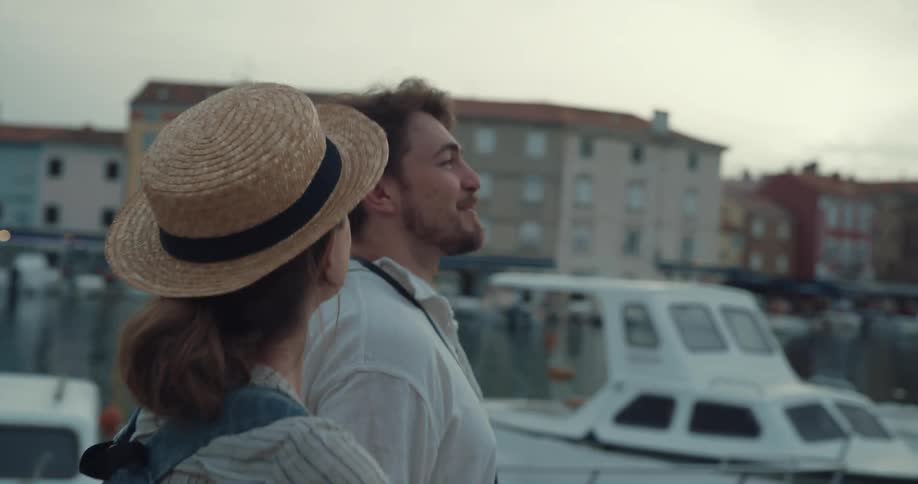 A man and woman gesture at their surroundings while walking through a marina in a town during the day