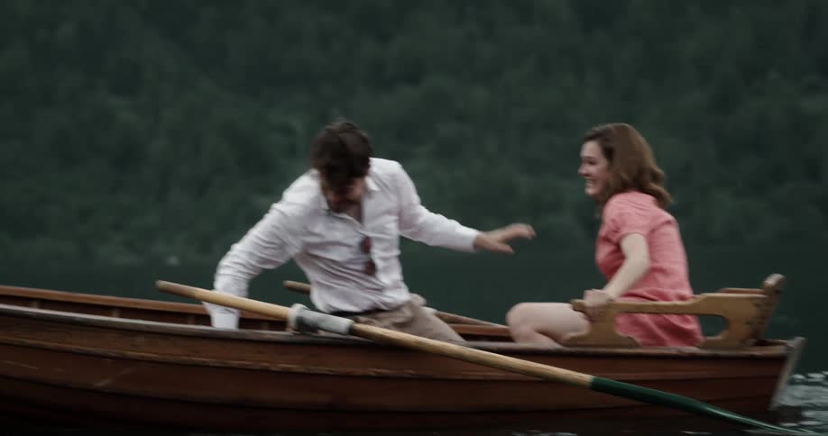 A man and woman splash water at each other while sitting in a boat on a lake during the day