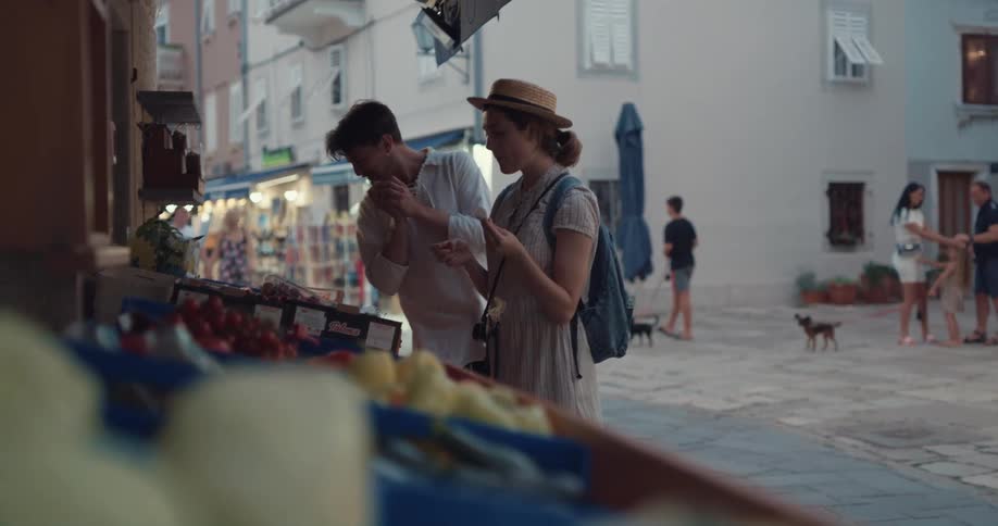 Couple browses through produce displayed outside shop as others walk past in background