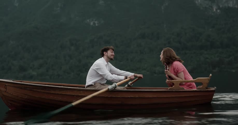 Man and woman sitting on a wooden canoe navigating a lake while the woman is playing a mini guitar and the man is rowing