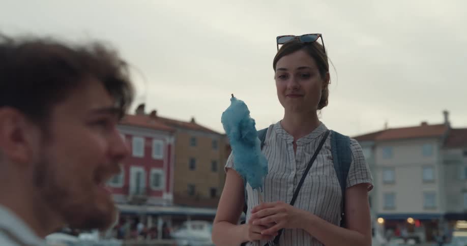 A woman holding cotton candy stands in front of a man playing a guitar on the sidewalk and drops some money in to his hat