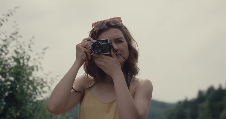 A woman standing at the edge of a forest holds a camera to her face and takes a picture