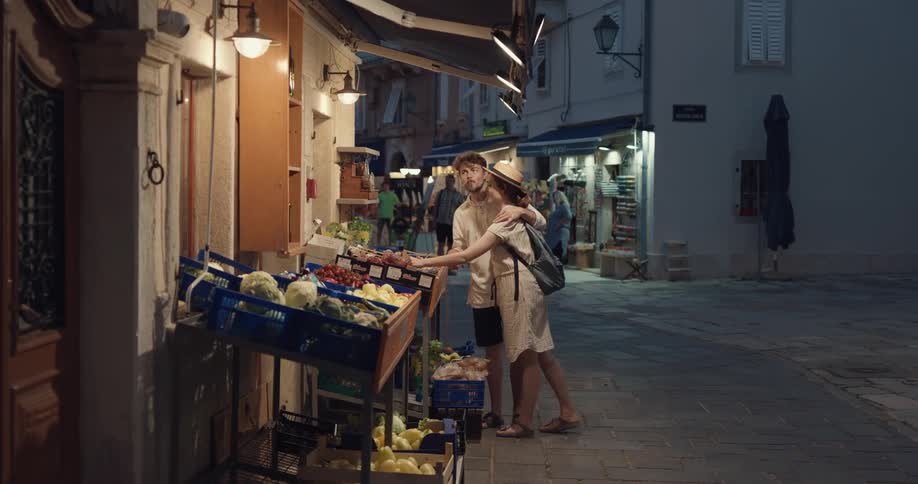 A man and woman that appear to be tourists stop at a street side market and look at food and then sample some food and walk away