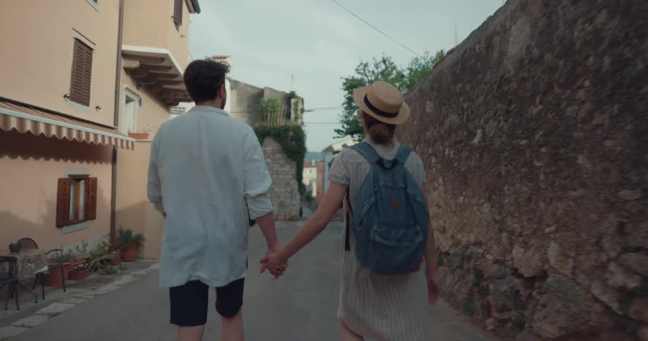 A man kisses a womans hand as they walk hand in hand down a road in a town during the day