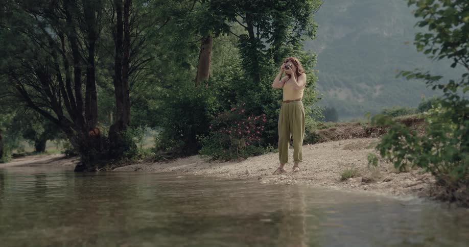 Woman standing next to the water in front of a lake taking pictures of the surroundings with her digital camera