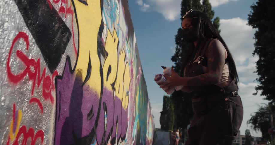 Black woman wearing a face mask doing graffiti in a wall at a public space in the middle of the day