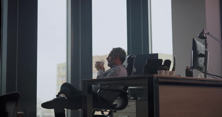 Man sitting in the chair facing the glass window on his office space relaxing and drinking a cup of coffee