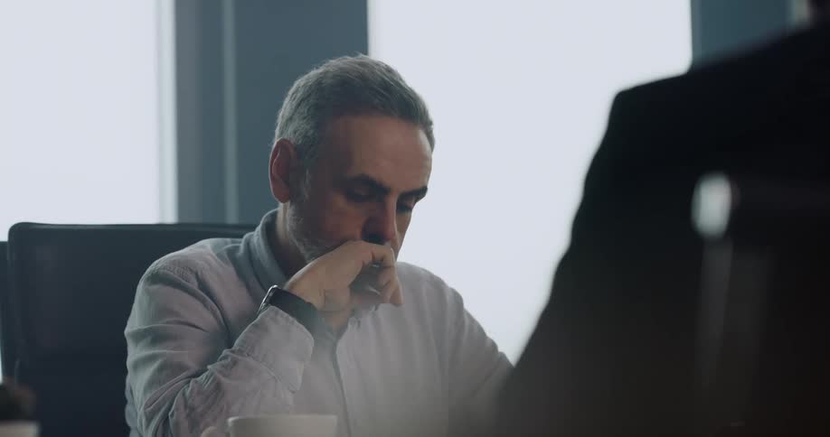 A man sitting at a table in an office checks his watch while listening to a person sitting across from him
