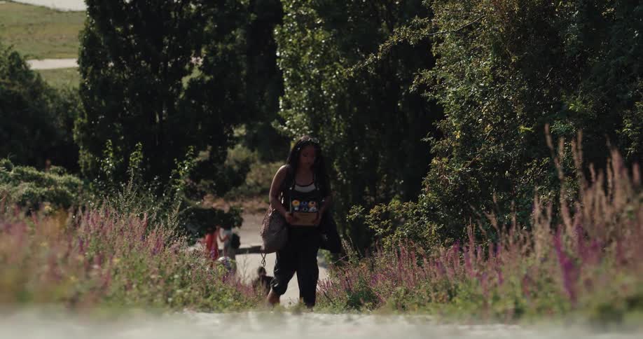 Black woman carrying a box filled with spray paint cans walking up a path during a sunny day