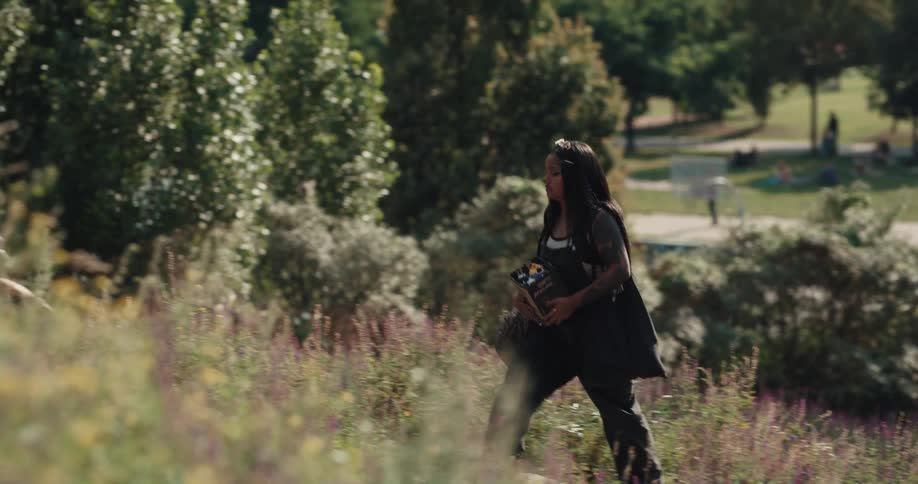 Black woman holding a box filled with spray paint cans walking up a path surrounded by vegetation at a public space during a sunny day
