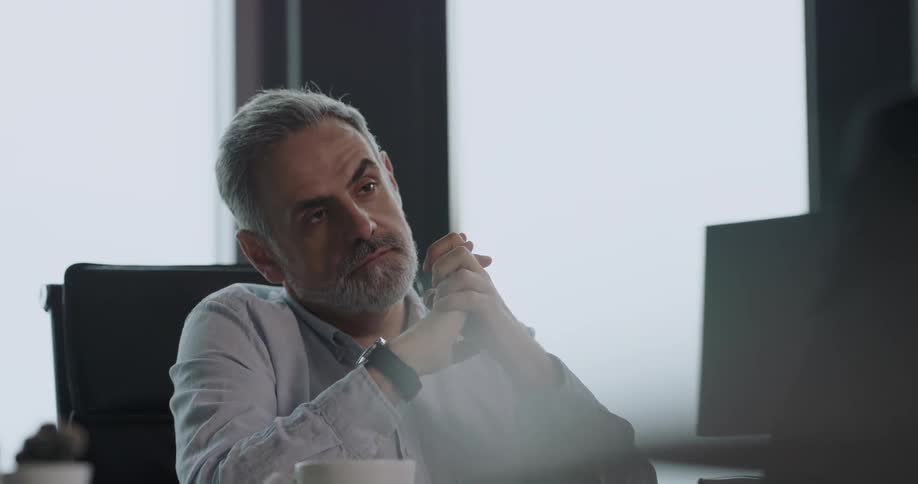 Bored businessman with beard and shirt looks at watch and listens to person in the office