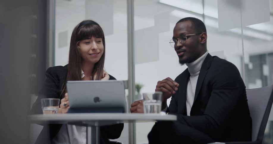 Man and woman sitting at the desk in an office room holding a video call on an ipad