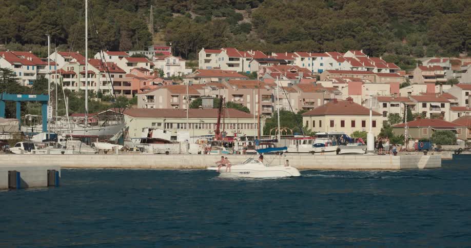 People sit on the front of a boat traveling in the harbor near a village as a bird flies overhead
