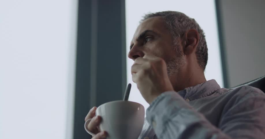 Middleaged man sitting on the office chair in front of a big window dipping a biscuit in the cup and eating it