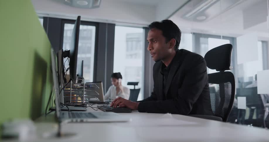 A man is in an office sitting at a desk typing on a computer keyboard