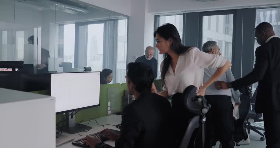 A woman points at a monitor and talks to a man while standing beside him as he sits before a computer at his desk