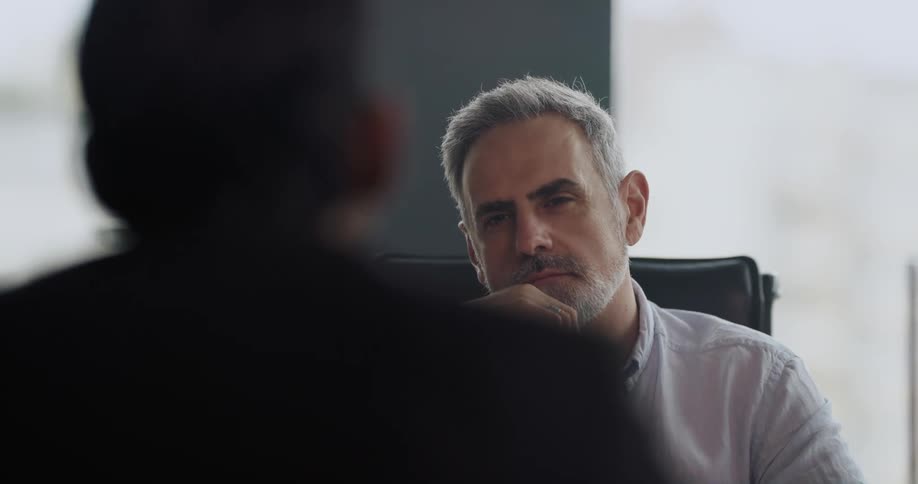 Man sitting at desk beckons and another man sits in chair in front of him as they talk