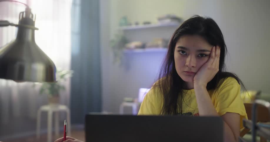 A girl is sitting at a table in front of a computer with a pen in her hand and the other hand on her face