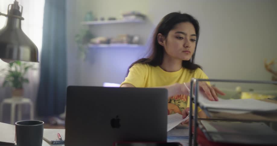 Young woman sitting at a desk in her room takes notes and organizes paperwork in front of a laptop