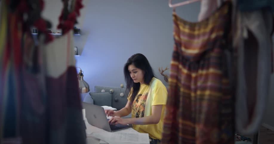 Tired and bored woman with straight hair and yellow and blue pajamas sits in chair and works on laptop in her room
