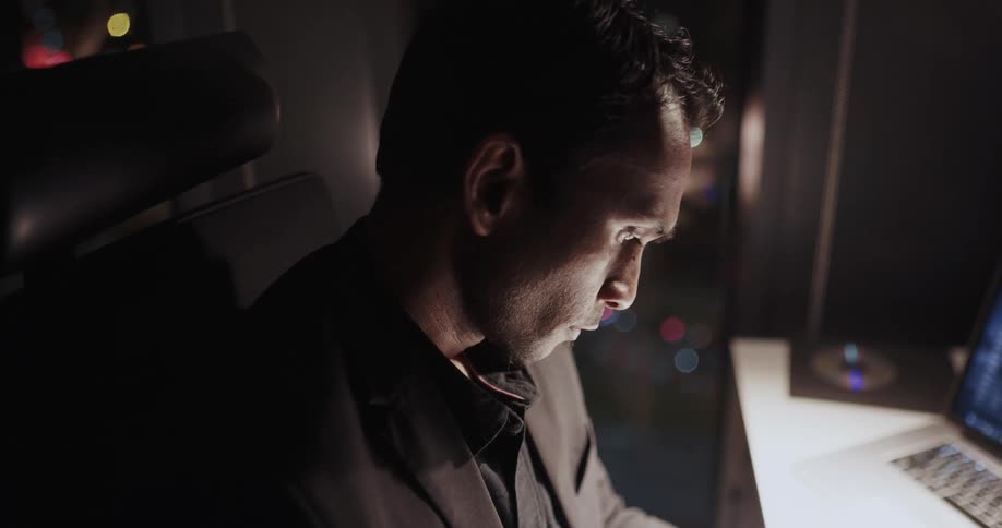 Business man sitting at the desk next to a big glass window on his office space at night