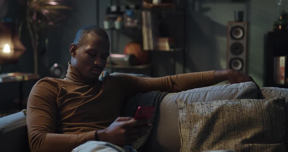 Man with brown turtleneck sweater and jeans sits on couch using smartphone in living room at home