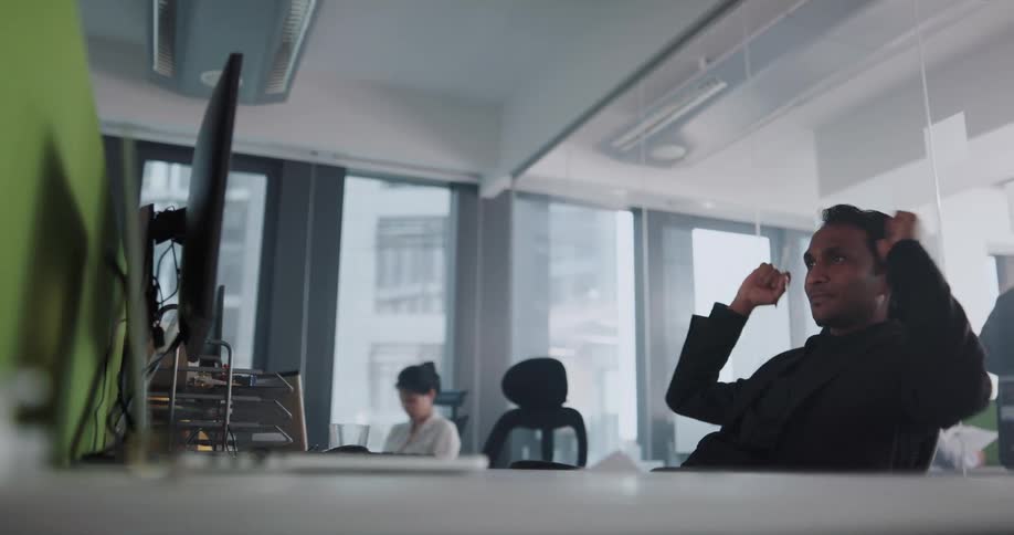 A man typing on a computer in a shared office raises his arms in celebration