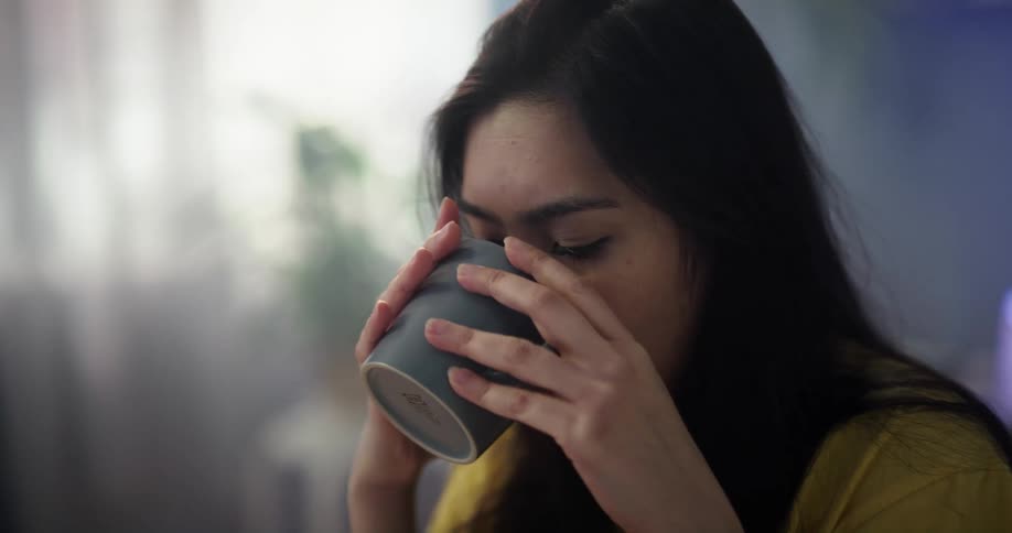 A tired young woman closes her laptop and drinks from her coffee mug before stretching her neck