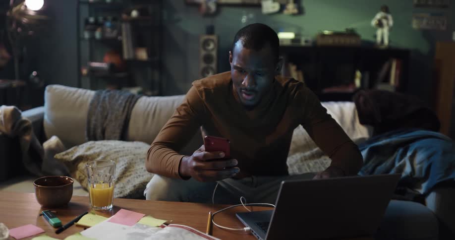 Man sits in living room with connected smartphone and laptop as he works then celebrates what he sees on the screens